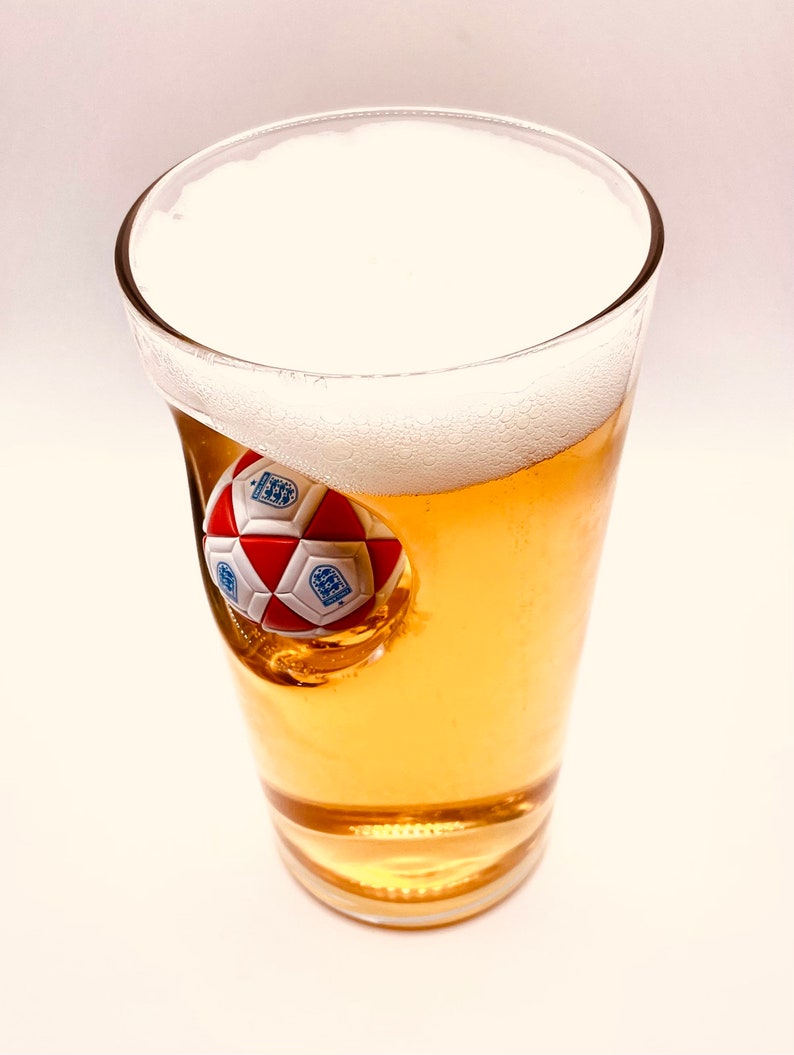 May include: A clear glass pint beer glass filled with golden liquid and a thick white foam head. A three-dimensional red and white soccer ball with blue England national team crests is embedded into the side of the glass, protruding inward.