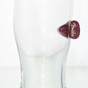 May include: A clear glass pint glass with a Fender guitar pick stuck to the side. The pick is red with gold lettering that says "Fender".