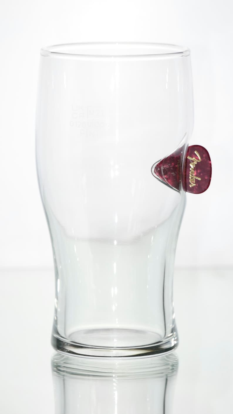 May include: A clear glass pint glass with a Fender guitar pick stuck to the side. The pick is red with gold lettering that says "Fender".