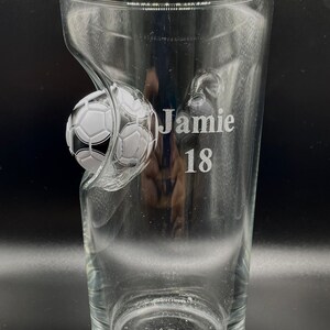 May include: A clear glass pint tumbler featuring an embedded black and white soccer ball design on the side. The glass is etched with the name Jamie and the number 18 in a clean, sans-serif font. The vessel has a smooth, transparent finish.