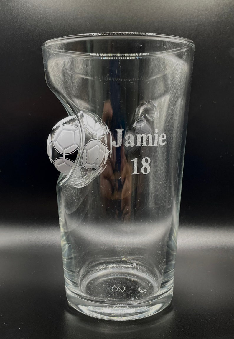 May include: A clear glass pint tumbler featuring an embedded black and white soccer ball design on the side. The glass is etched with the name Jamie and the number 18 in a clean, sans-serif font. The vessel has a smooth, transparent finish.
