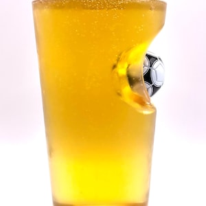 May include: A clear glass pint glass filled with a golden yellow liquid, featuring a unique indentation on one side that contains a small, black and white soccer ball embedded into the glass structure, creating a distinct three-dimensional effect.