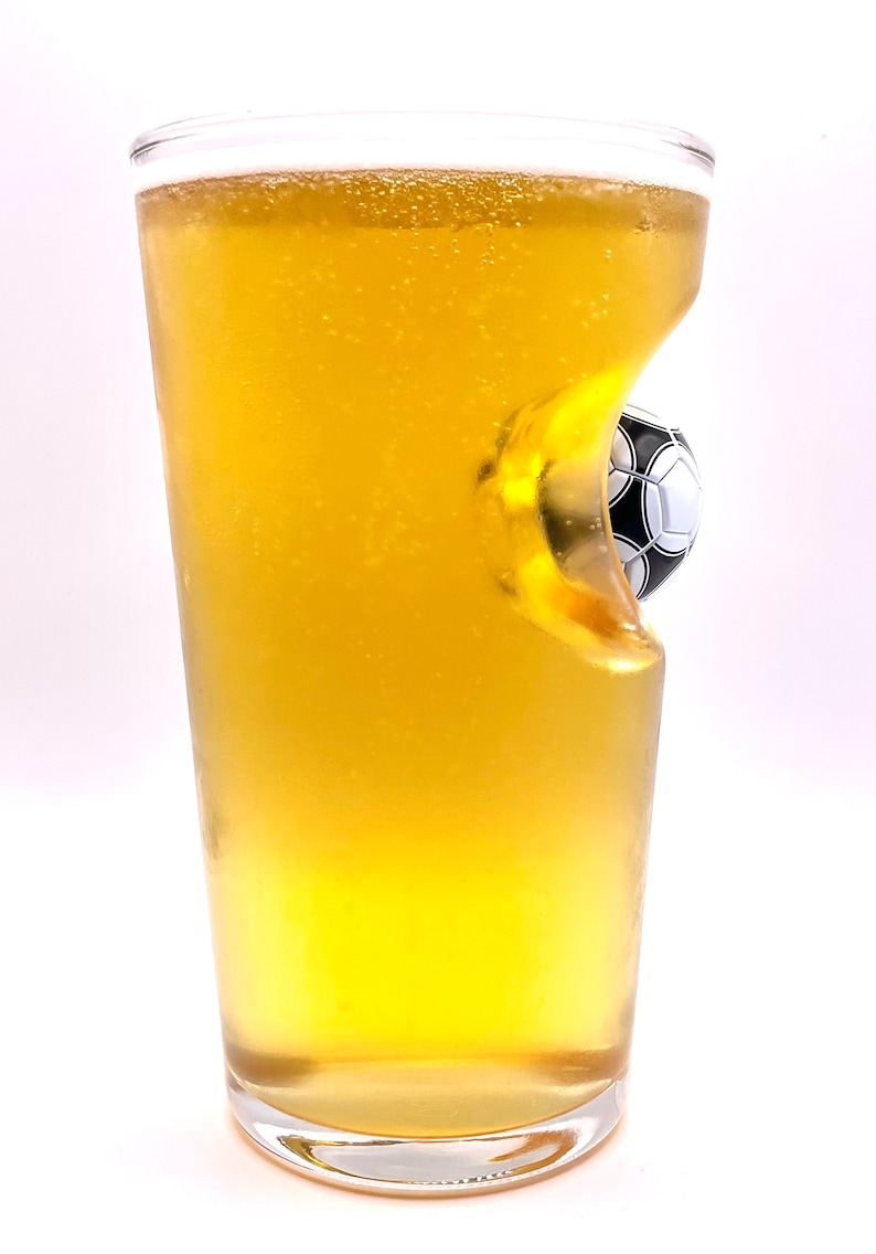 May include: A clear glass pint glass filled with a golden yellow liquid, featuring a unique indentation on one side that contains a small, black and white soccer ball embedded into the glass structure, creating a distinct three-dimensional effect.
