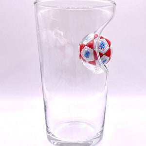 May include: A clear glass tumbler featuring a molded indentation on one side that holds a miniature soccer ball. The ball is patterned with red and white pentagons and hexagons, and it displays the blue and white England national team crest.