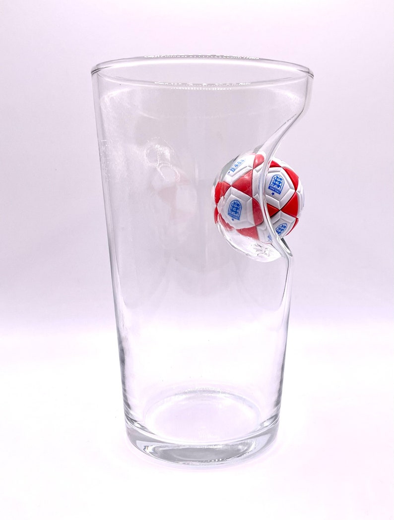 May include: A clear glass tumbler featuring a molded indentation on one side that holds a miniature soccer ball. The ball is patterned with red and white pentagons and hexagons, and it displays the blue and white England national team crest.