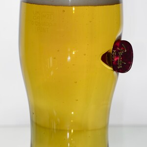 May include: A clear glass pint glass filled with a light yellow beer. The glass has a small, red Fender guitar pick stuck to the side. The glass has markings that read "UK CP M21 0.125 UK PINT".