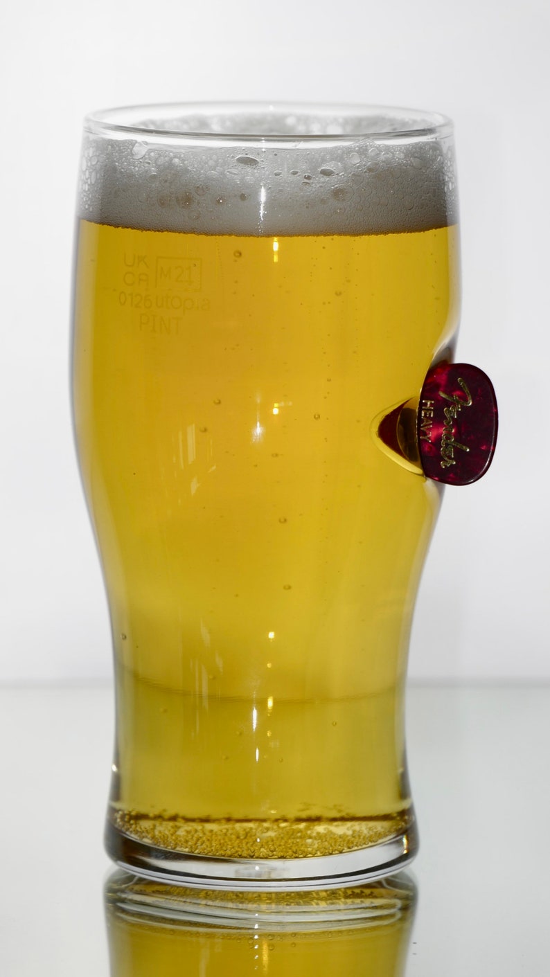 May include: A clear glass pint glass filled with a light yellow beer. The glass has a small, red Fender guitar pick stuck to the side. The glass has markings that read "UK CP M21 0.125 UK PINT".