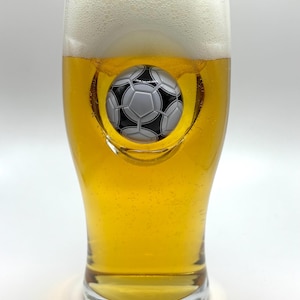 May include: A clear glass beer pint filled with golden liquid and a thick white foam head. The glass features a three-dimensional, black and white soccer ball embedded into the center of the glass wall, creating a concave indentation.