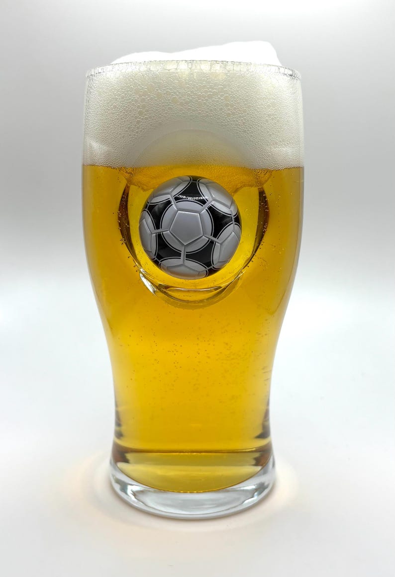 May include: A clear glass beer pint filled with golden liquid and a thick white foam head. The glass features a three-dimensional, black and white soccer ball embedded into the center of the glass wall, creating a concave indentation.