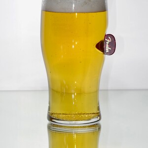 May include: A clear glass pint glass filled with a light yellow beer, with a white head of foam. A red Fender guitar pick is attached to the side of the glass.