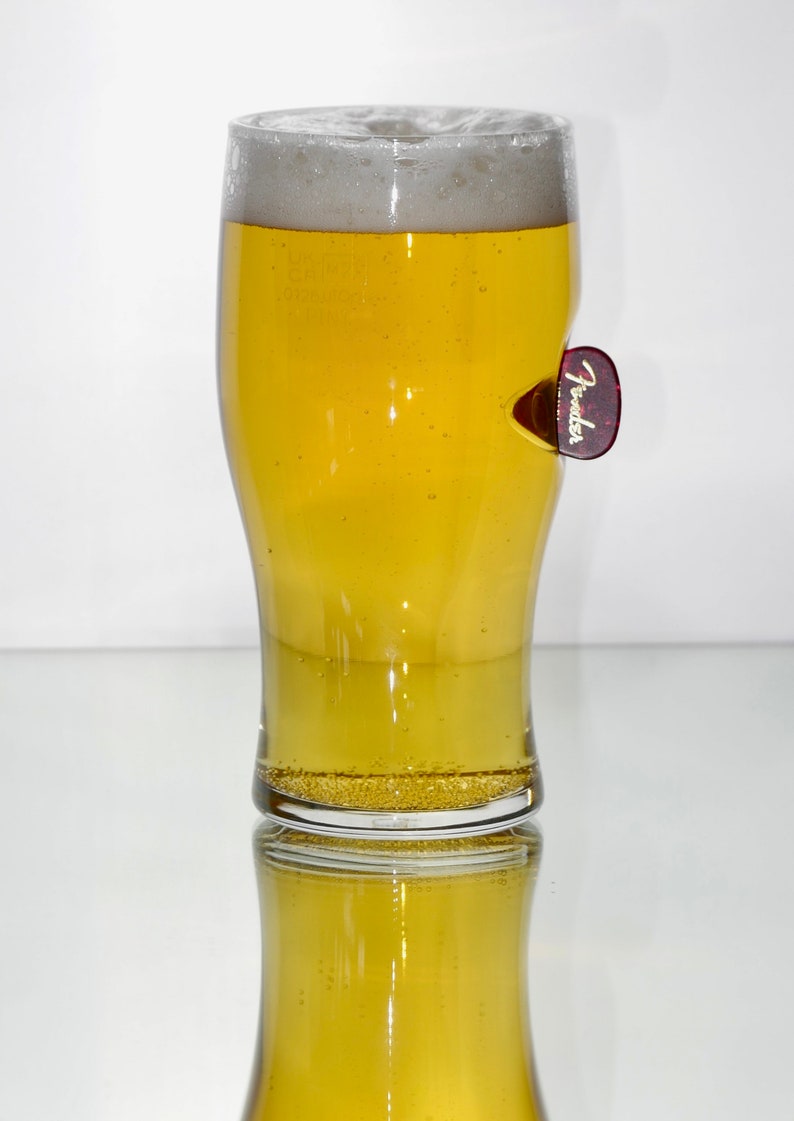 May include: A clear glass pint glass filled with a light yellow beer, with a white head of foam. A red Fender guitar pick is attached to the side of the glass.
