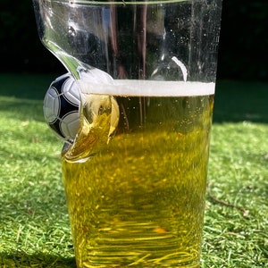 May include: A clear glass pint glass filled with a golden yellow liquid and white foam, featuring a three-dimensional black and white soccer ball embedded into the side of the glass wall, creating a distinct indentation in the clear glass structure.
