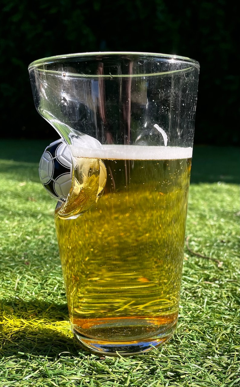 May include: A clear glass pint glass filled with a golden yellow liquid and white foam, featuring a three-dimensional black and white soccer ball embedded into the side of the glass wall, creating a distinct indentation in the clear glass structure.