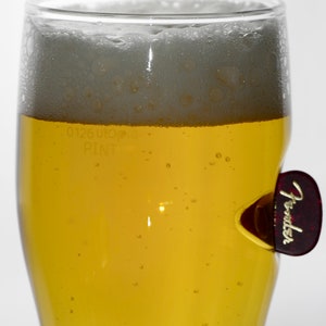 May include: A glass of light yellow beer with a white head of foam. The glass has a sticker on the side that says "Fender".