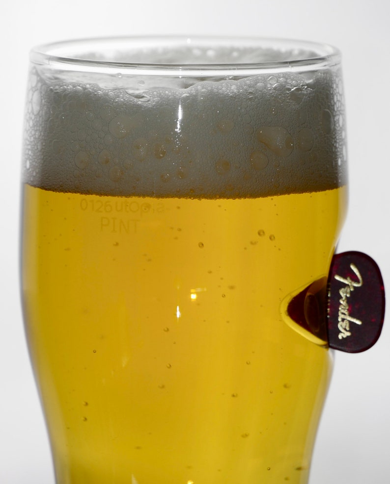 May include: A glass of light yellow beer with a white head of foam. The glass has a sticker on the side that says "Fender".