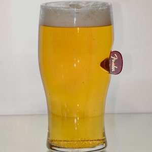 May include: A clear glass beer mug with a frosted rim filled with a light yellow beer. A red guitar pick with the word "Fender" in gold letters is attached to the side of the mug.