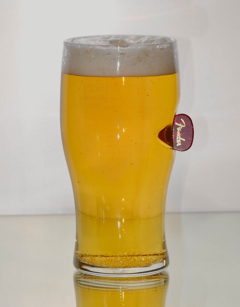 May include: A clear glass beer mug with a frosted rim filled with a light yellow beer. A red guitar pick with the word "Fender" in gold letters is attached to the side of the mug.