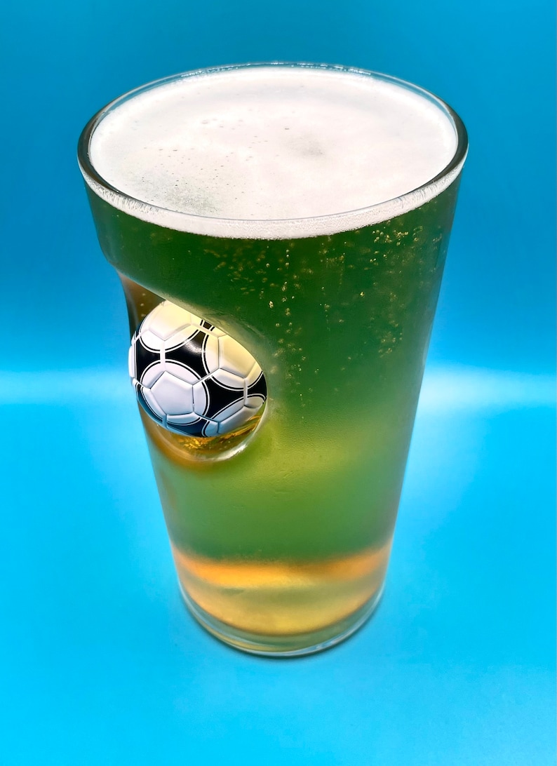 May include: A clear glass pint glass filled with a green liquid and a white foam head. The glass features a molded, three-dimensional black and white soccer ball embedded into the side, creating a concave indentation within the glass structure.