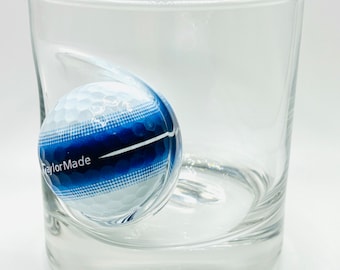 Könnte beinhalten: Ein durchsichtiger Glasbecher mit einem blauen und weißen Golfball-Design. Der Golfball ist teilweise im Glas versunken und trägt den Schriftzug "TaylorMade".