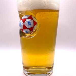 May include: A clear glass pint beer mug filled with golden lager and a thick white foam head. A three-dimensional red and white soccer ball with the blue England national team crest is embedded into the side of the glass, creating a concave indentation.