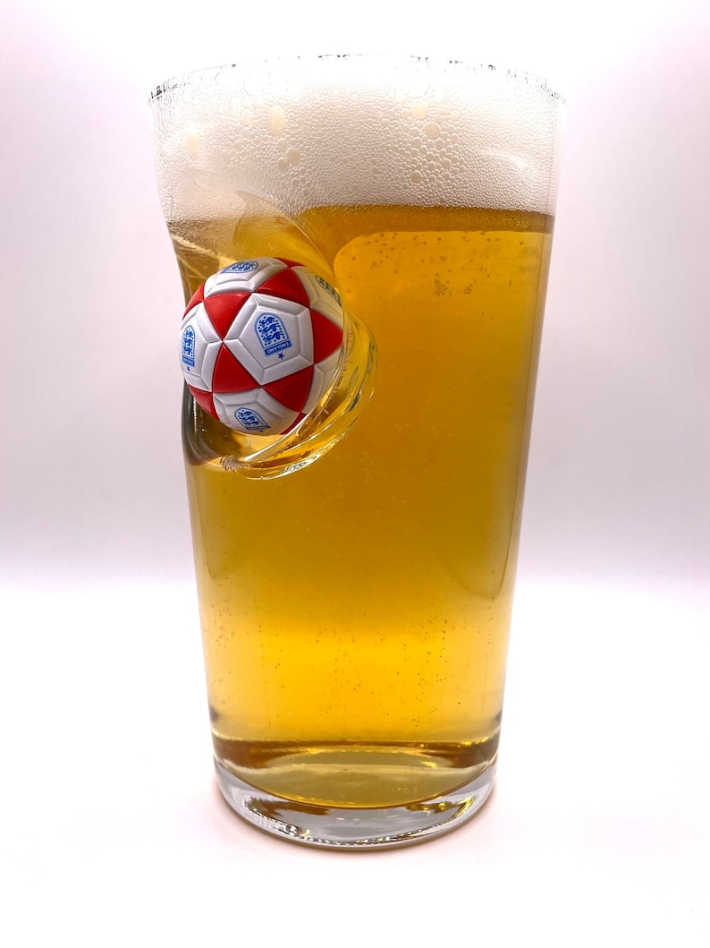 May include: A clear glass pint beer mug filled with golden lager and a thick white foam head. A three-dimensional red and white soccer ball with the blue England national team crest is embedded into the side of the glass, creating a concave indentation.
