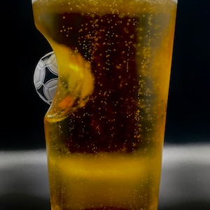 May include: A clear glass pint beer glass filled with a golden carbonated beverage. The side of the glass features a molded indentation shaped like a black and white soccer ball, creating a unique visual effect as the liquid fills the glass.