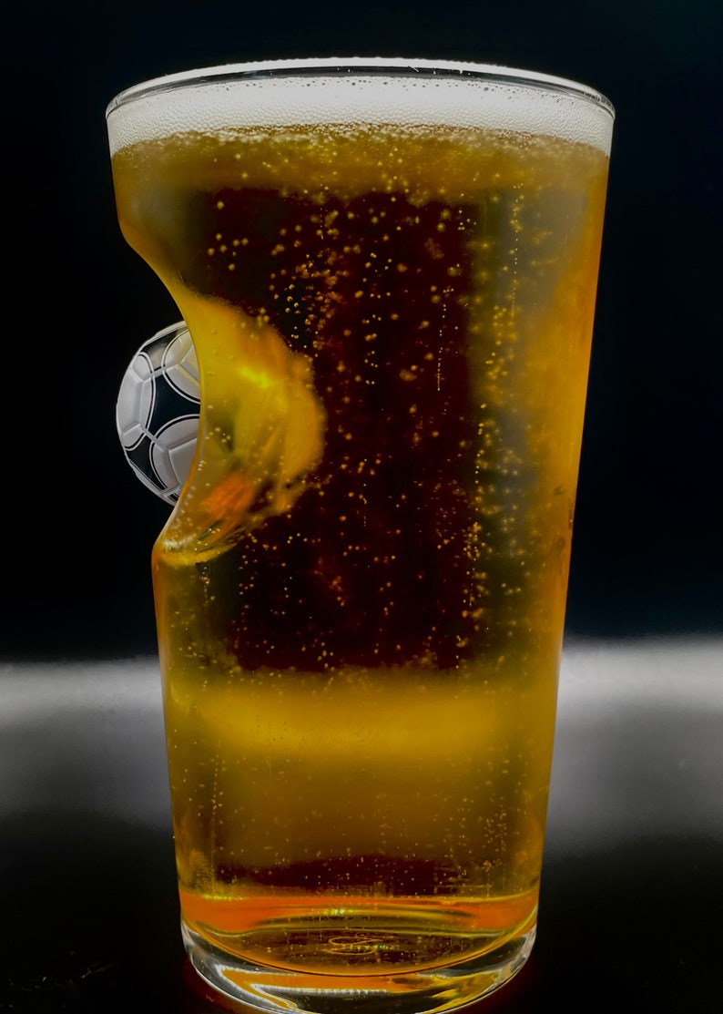 May include: A clear glass pint beer glass filled with a golden carbonated beverage. The side of the glass features a molded indentation shaped like a black and white soccer ball, creating a unique visual effect as the liquid fills the glass.