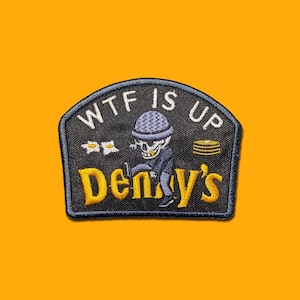 May include: Embroidered patch with the text "WTF IS UP" above "Denny's". The patch features a cartoon skeleton wearing a beanie and a jacket, with pancakes and eggs. The patch is black with yellow and white text.