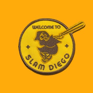 May include: A yellow and brown embroidered patch featuring a skeleton figure swinging a baseball bat. The patch reads "Welcome to Slam Diego".