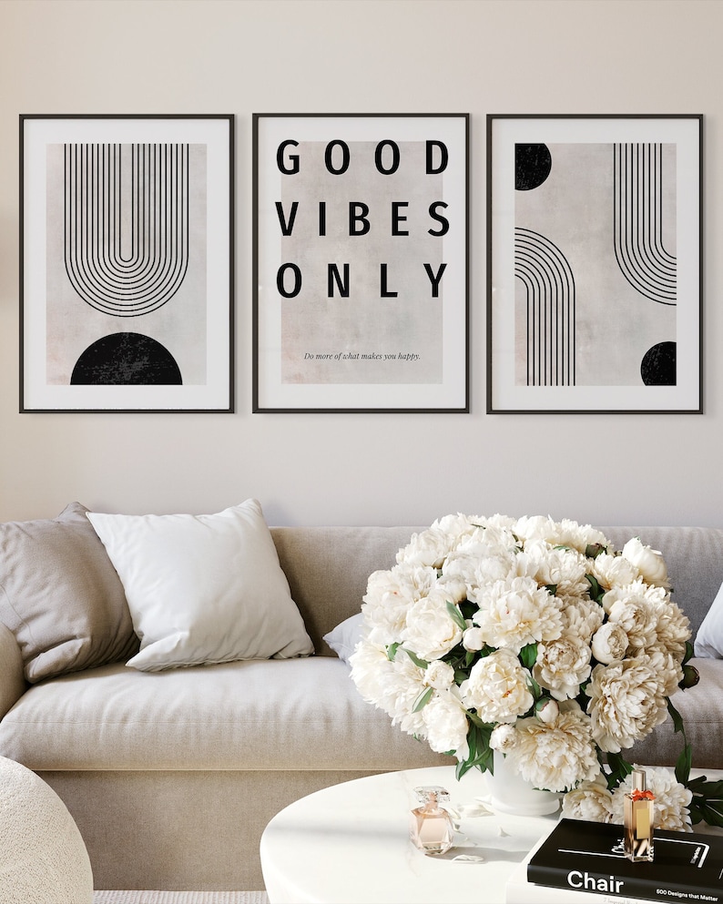 May include: Three framed art prints with black frames. The prints feature abstract designs in black and beige. The center print reads "GOOD VIBES ONLY" in large black letters. A sofa and coffee table with a floral arrangement are in the foreground.