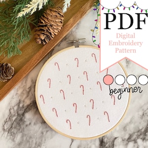 Puede incluir: Un aro de bordado blanco con un patrón de bastones de caramelo rojos. El aro está sobre una superficie de mármol con una bandeja de madera y ramas de pino en el fondo. El texto "PDF Digital Embroidery Pattern" y "beginner" es visible en la imagen.