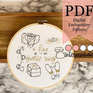 Puede incluir: Un aro de bordado blanco con un diseño de bordado dorado de una rosa, una cara de gato, una tetera, guantes y una caja de regalo. El texto "a few of my favorite things" también está bordado en oro. El aro está sobre una superficie de madera. Este es un patrón de bordado digital para niveles intermedios.