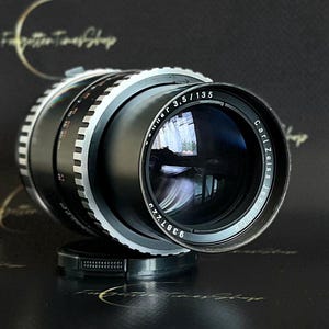 May include: A black Carl Zeiss Jena 135mm f/3.5 lens with a silver ring around the barrel. The lens is in focus and the aperture is open.