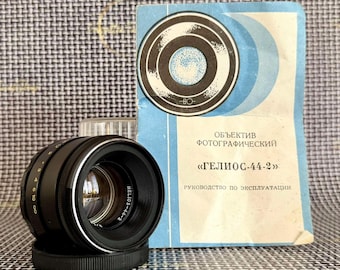 May include: A black Helios-44-2 camera lens with a silver ring and focus markings, positioned next to a light blue instruction manual featuring a circular graphic and the text "ОБЪЕКТИВ ФОТОГРАФИЧЕСКИЙ ГЕЛИОС-44-2" printed in black Cyrillic lettering.