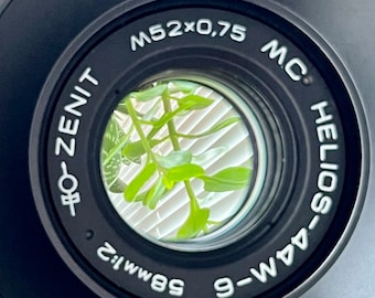 May include: Black camera lens with the text "M52x0,75 MC HELIOS-44M-6" and "58mm:2". The lens reflects a view of green plants and white blinds. The lens is a photography accessory.