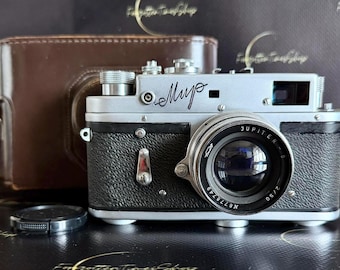 May include: A silver and black rangefinder camera with a Jupiter-8 2/50 lens and the Cyrillic text "Mup" on the front. The camera is accompanied by a brown leather case and a separate black lens cap, featuring metallic dials and a textured grip.