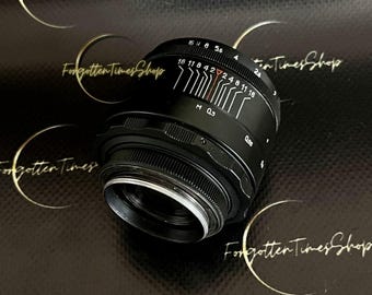 May include: A black camera lens with white markings and a focus ring. The lens has a series of numbers and indicators, including "16 11 8 4 2 2.4 8 11 16". The text "Forgotten Times Shop" is visible in the background.