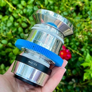 May include: A silver camera lens with a blue gear-like ring and black accents. The lens has a shiny, metallic finish and features the text "M42-FX". The background is blurred green foliage with a red flower.