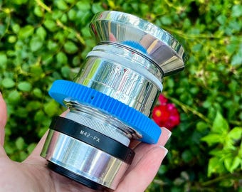 May include: A silver camera lens with a blue gear-like ring and black accents. The lens has a shiny, metallic finish and features the text "M42-FX". The background is blurred green foliage with a red flower.