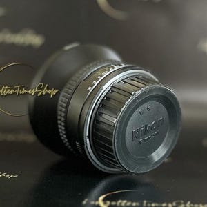 May include: A black Nikon camera lens with a silver ring and a black lens cap. The lens cap has the Nikon logo and the text "F-mount" printed on it.