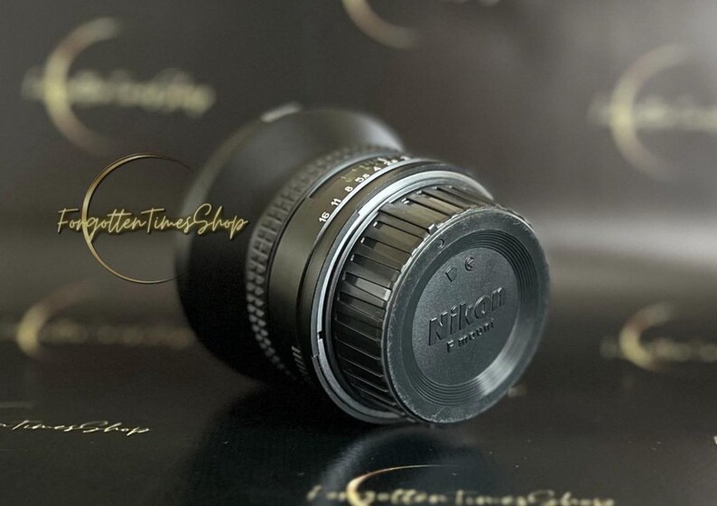 May include: A black Nikon camera lens with a silver ring and a black lens cap. The lens cap has the Nikon logo and the text "F-mount" printed on it.