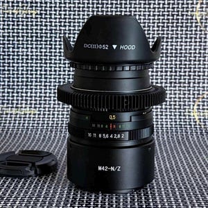 May include: A black camera lens with a lens hood and a lens cap. The lens has markings for aperture and focus, with the text "DC(II)Φ52 HOOD" and "M42-N/Z" visible. The lens is on a patterned surface.