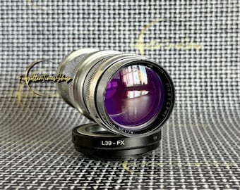 May include: A silver-toned camera lens with a purple-tinted front glass element, featuring Cyrillic text and the markings 4/135 on the front rim. The lens is attached to a black circular adapter ring clearly labeled with the text L39 - FX.