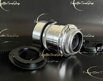 May include: A silver camera lens with a chrome finish and a black lens cap. The lens has a depth of field scale and aperture markings. The lens is sitting on a black surface.