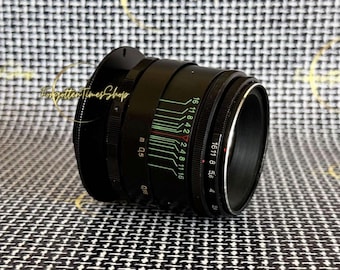 May include: Black camera lens with white and green markings, including f-stop numbers. The lens has a focus ring and aperture ring. The lens is set against a black and white checkered background.