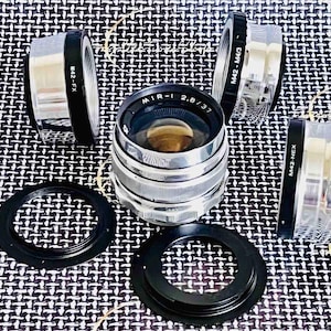 May include: A collection of camera lenses and adapters. The central lens is silver with the text "MIR-1 2.8/37". Surrounding it are silver and black adapters labeled "M42-FX", "M42-M4/3", and "M42-NEX". Two black rings are also included.