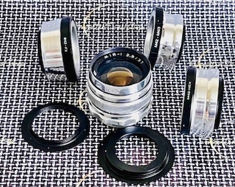 May include: A collection of camera lenses and adapters. The central lens is silver with the text "MIR-1 2.8/37". Surrounding it are silver and black adapters labeled "M42-FX", "M42-M4/3", and "M42-NEX". Two black rings are also included.