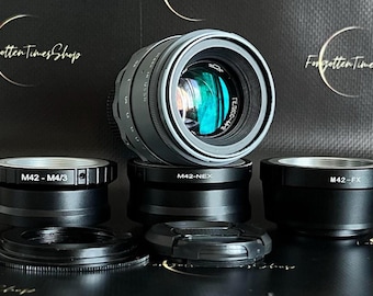 May include: A silver camera lens with a black lens cap and three black adapter rings labeled "M42 - M4/3", "M42-NEX", and "M42-FX".