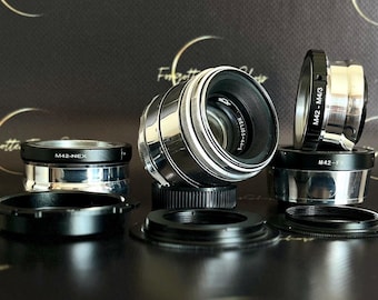 May include: A collection of camera lenses and adapters, including a silver and black Helios-44-2 lens and several M42 adapters. The items are arranged on a dark surface, with text on the adapters indicating their compatibility.