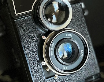 May include: A black and silver 166B LOMO camera with a lens that says "LOMO" and "MADE IN USSR" on it. The camera has a black textured body and a silver lens cap.